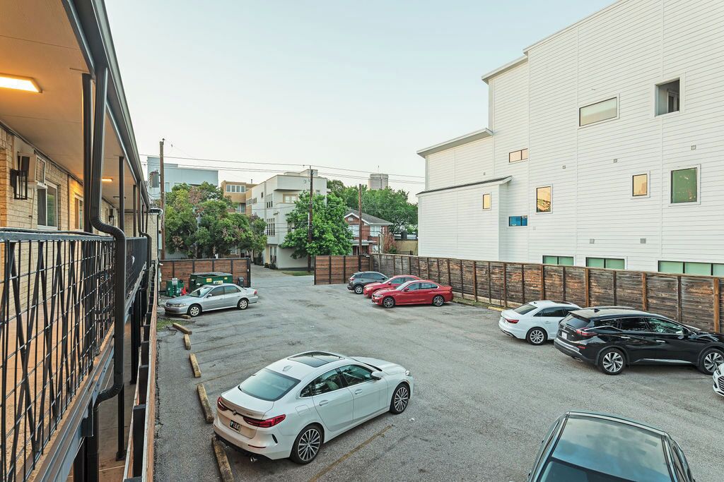 Ein Parkplatz mit mehreren Autos, ein Gebäude mit weißer Fassade und ein von Bäumen gesäumtes, eingezäuntes Areal.