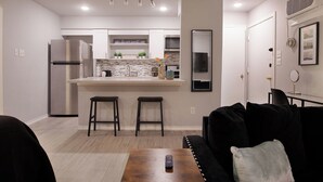 A modern kitchen with a central island, bar stools, a refrigerator, and a dining area with a black sofa. - Montrose Dazzle Studio, Near Med Center (Houston)