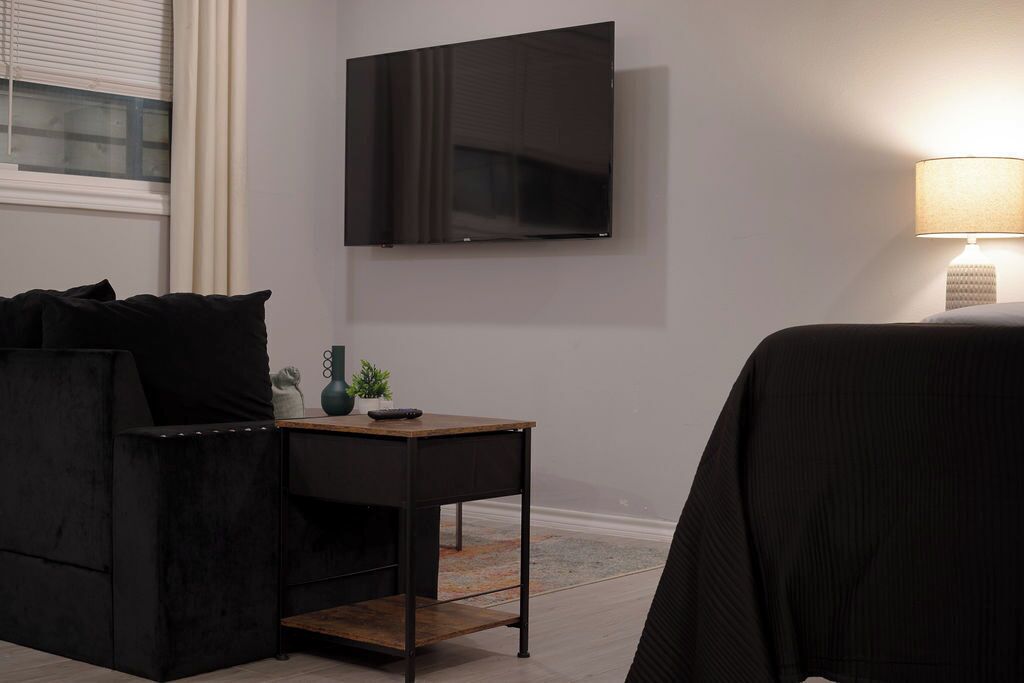 A modern room with a flat-screen TV mounted on the wall, a small wooden side table with a plant, and a lamp on the right.