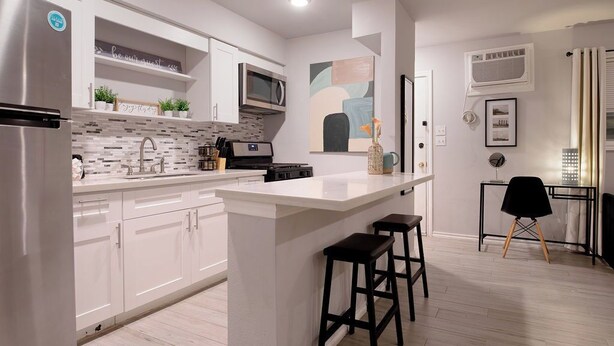A modern kitchen with white cabinetry, a central island with stools, a microwave, and a refrigerator. - Montrose Dazzle Studio, Near Med Center (Houston)
