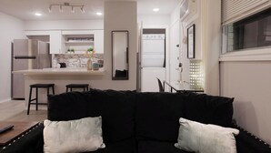 A modern living room with a black sofa, a small kitchen area with a refrigerator, oven, and bar stools. - Montrose Dazzle Studio, Near Med Center (Houston)