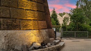 Un muro de piedra con estructura metálica, un área pavimentada con cercado de metal y árboles al fondo.
