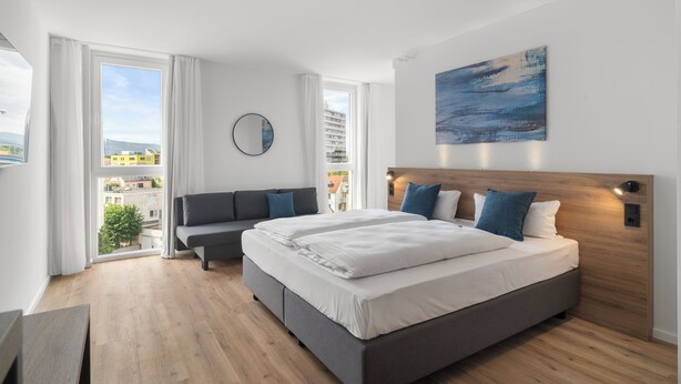 Un dormitorio moderno con una cama grande, un sofá, vistas a la ciudad y un cuadro en la pared.