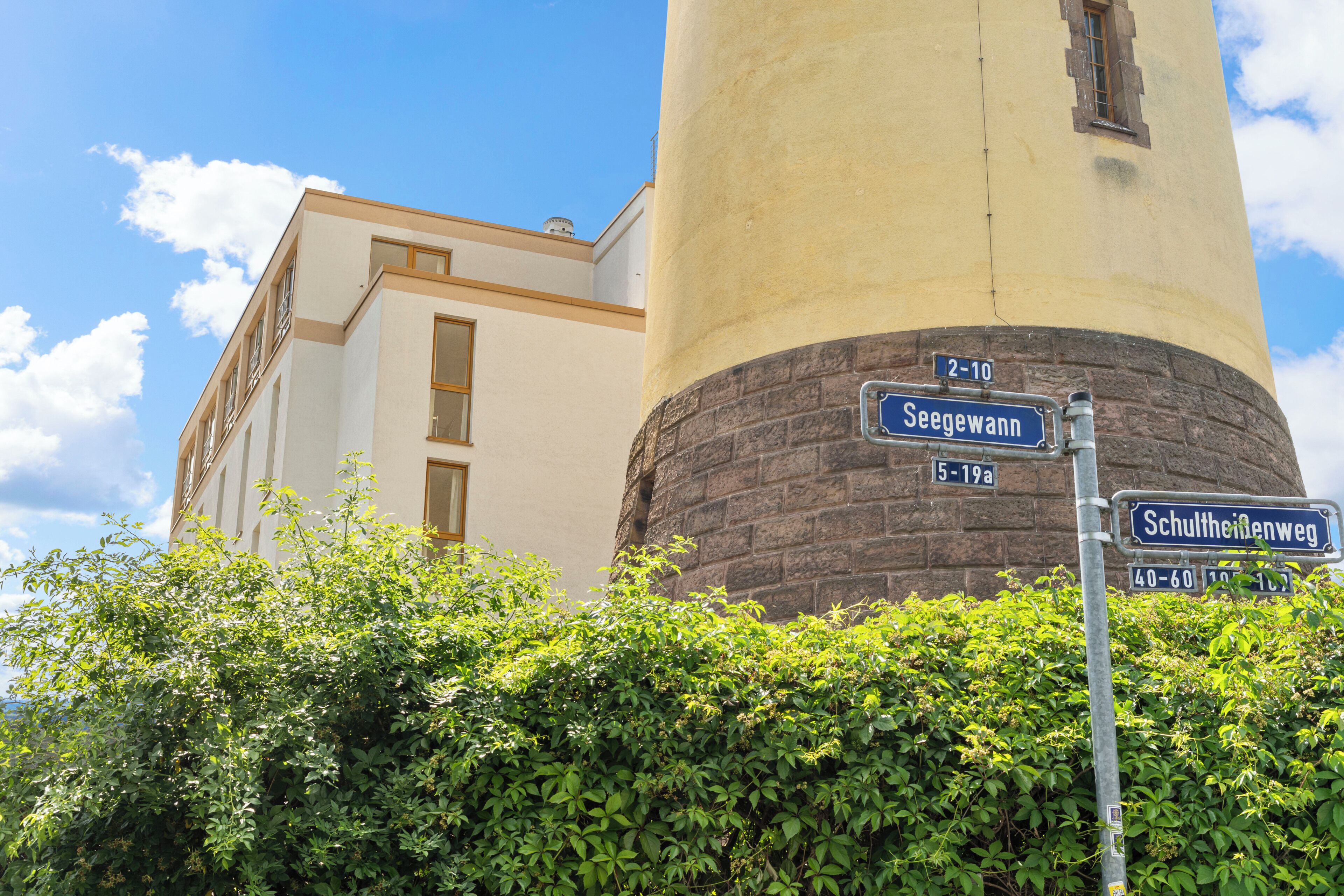 A building with a cylindrical tower, street signs for 'Seegwahn' and 'Schultheinenweg,' and a hedge in the foreground.