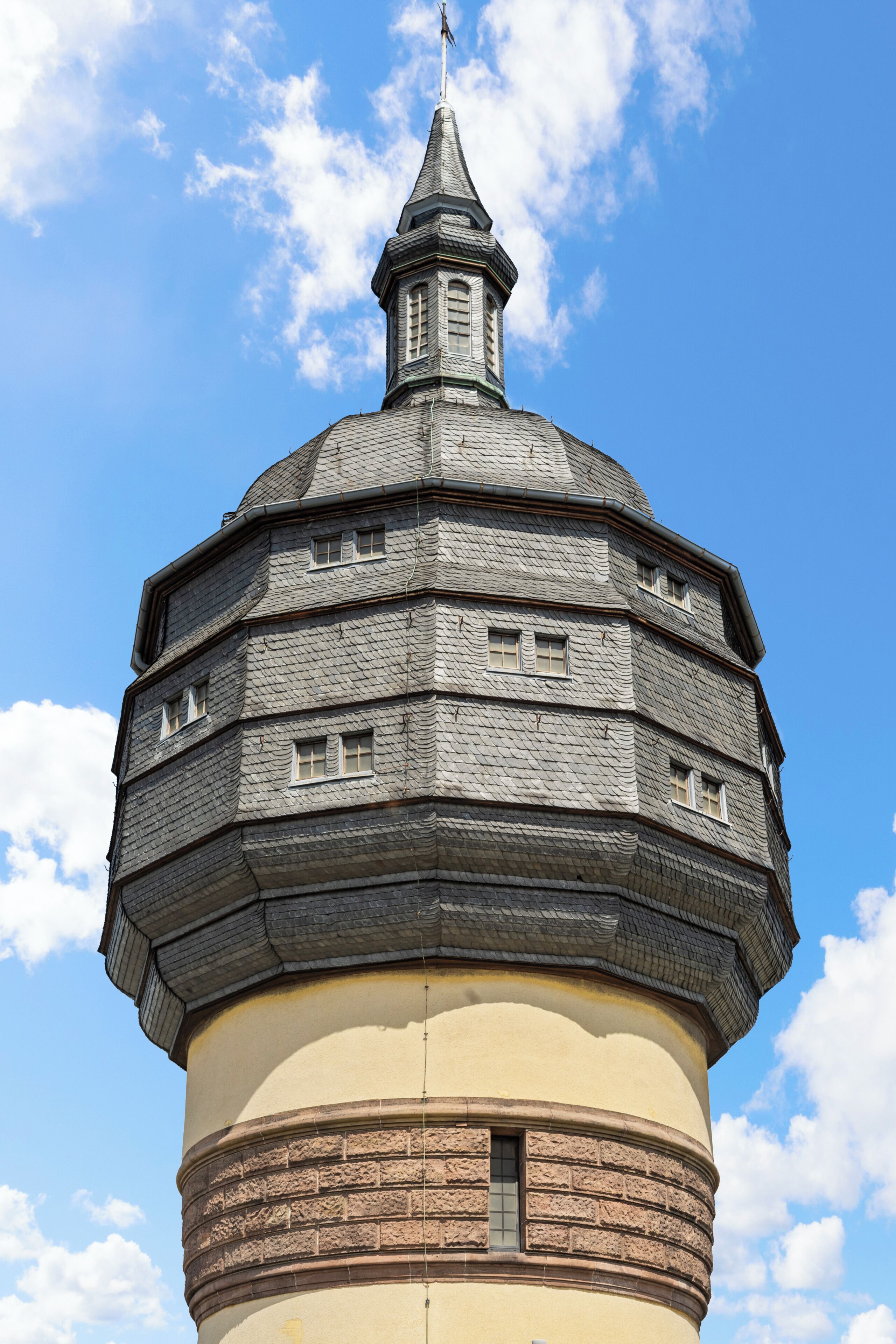 A tower with a conical roof and a spire, featuring multiple windows and a brick base.