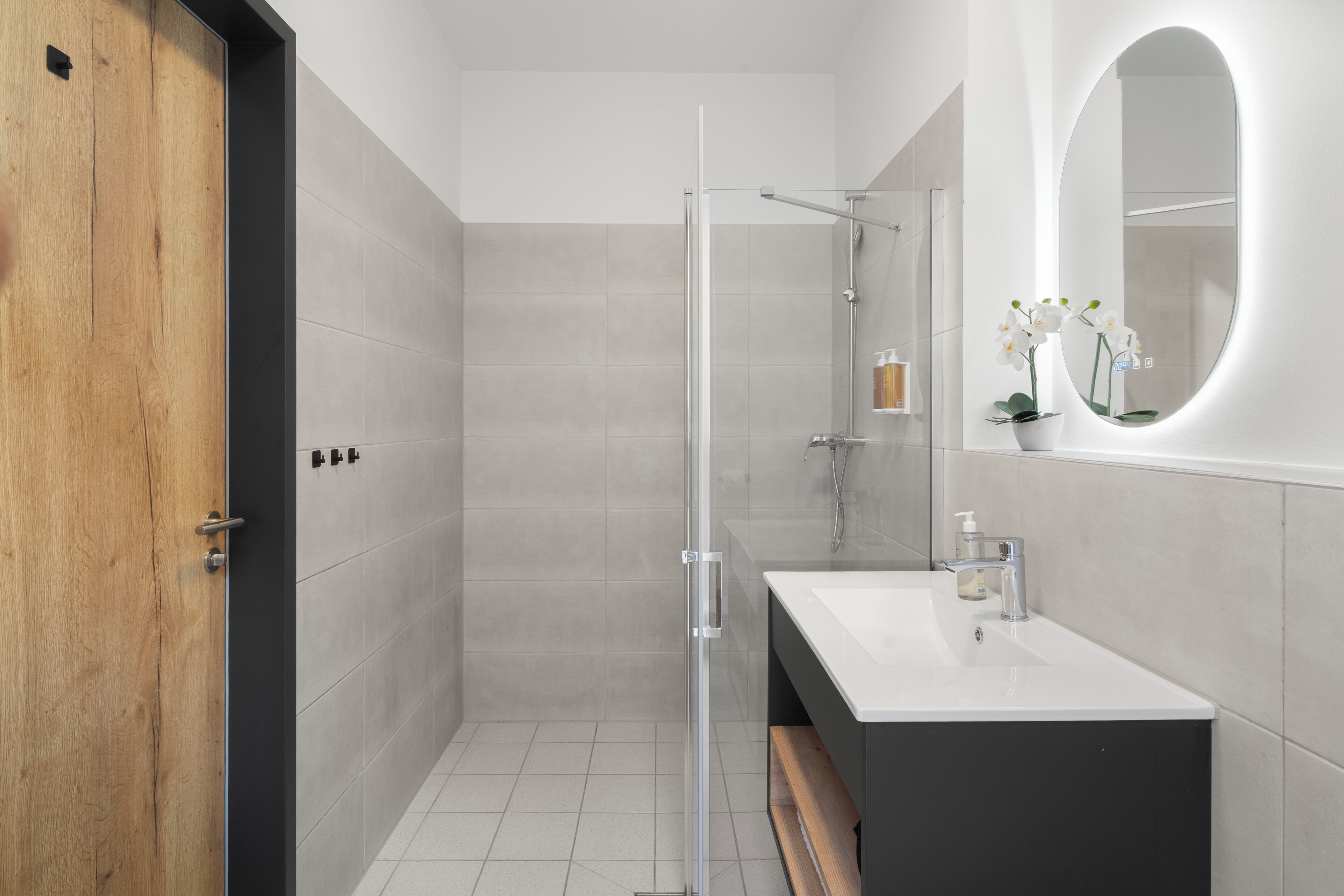 A modern bathroom with a glass shower enclosure, a wooden door, a white sink, and a mirror with an oval frame.