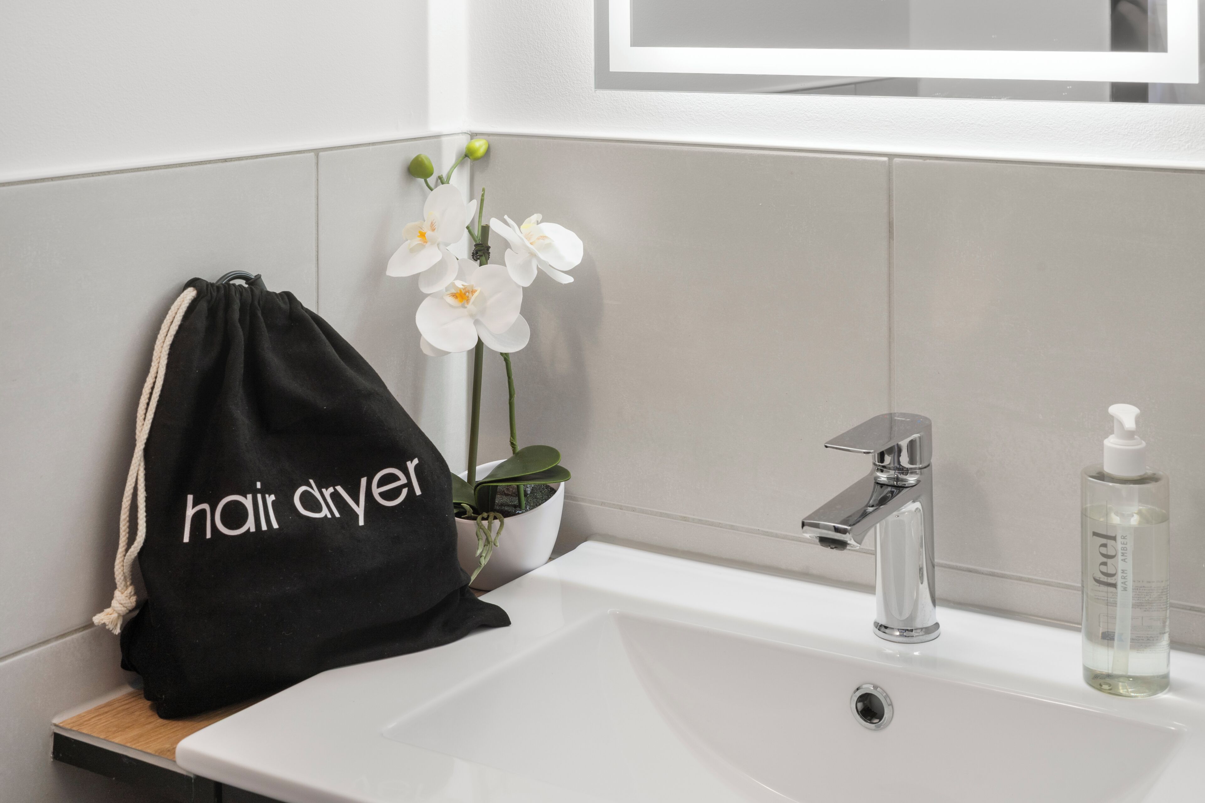 Un baño con un lavabo blanco, un espejo encima, una bolsa negra con el texto 'secador de pelo', una orquídea en maceta y una botella de jabón líquido.