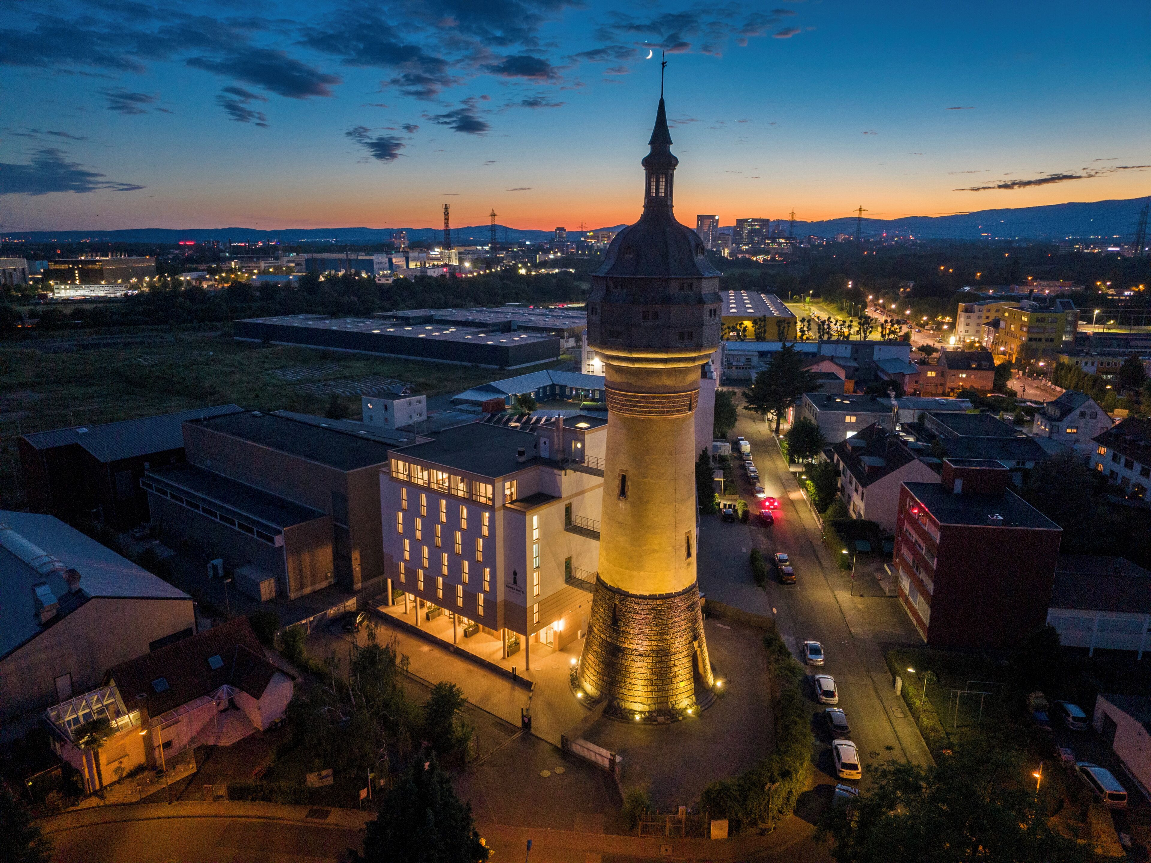 A historic tower with a clock, surrounded by modern buildings and a parking area.