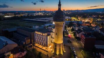 A historic tower with a clock, surrounded by modern buildings and a parking area.