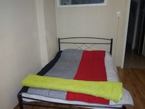 Una cama individual con estructura metálica, una pared lisa y una puerta al fondo.