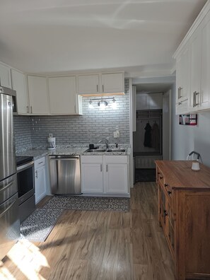 A modern kitchen with stainless steel appliances, white cabinetry, a gray backsplash, and a wooden dining table. - Near Airport cozy 2 bedroom  (Anchorage)