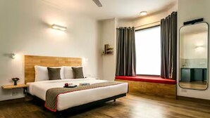 A hotel room with a wooden floor, a bed with a headboard, a bedside table, a window with curtains, and a mirror. - Classic Hotel Dev Haveli (Port Blair)