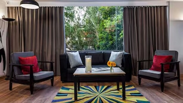 A room with a black sofa, two armchairs, a coffee table, a colorful rug, and a view of greenery through large windows. - Classic Hotel Dev Haveli (Port Blair)