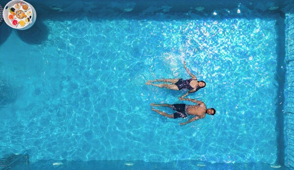 Une piscine rectangulaire à l’eau d’un bleu limpide, où flottent deux personnes, ainsi qu’un plateau flottant sur lequel se trouvent des aliments.