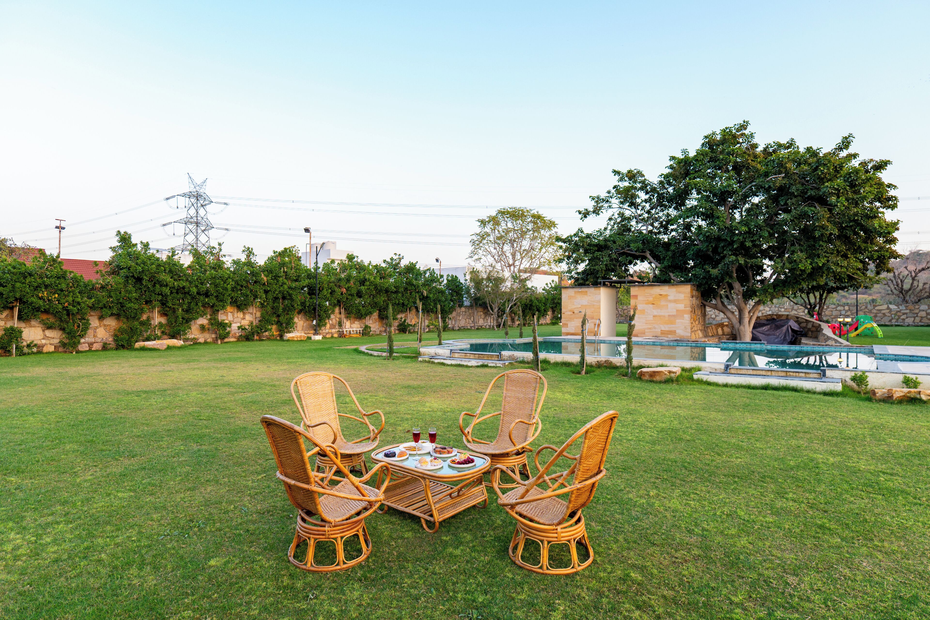 A grassy outdoor area with a table and four wicker chairs, a stone wall, and a swimming pool in the background.