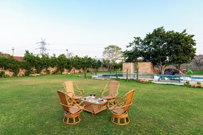 A grassy outdoor area with a table and four wicker chairs, a stone wall, and a swimming pool in the background.