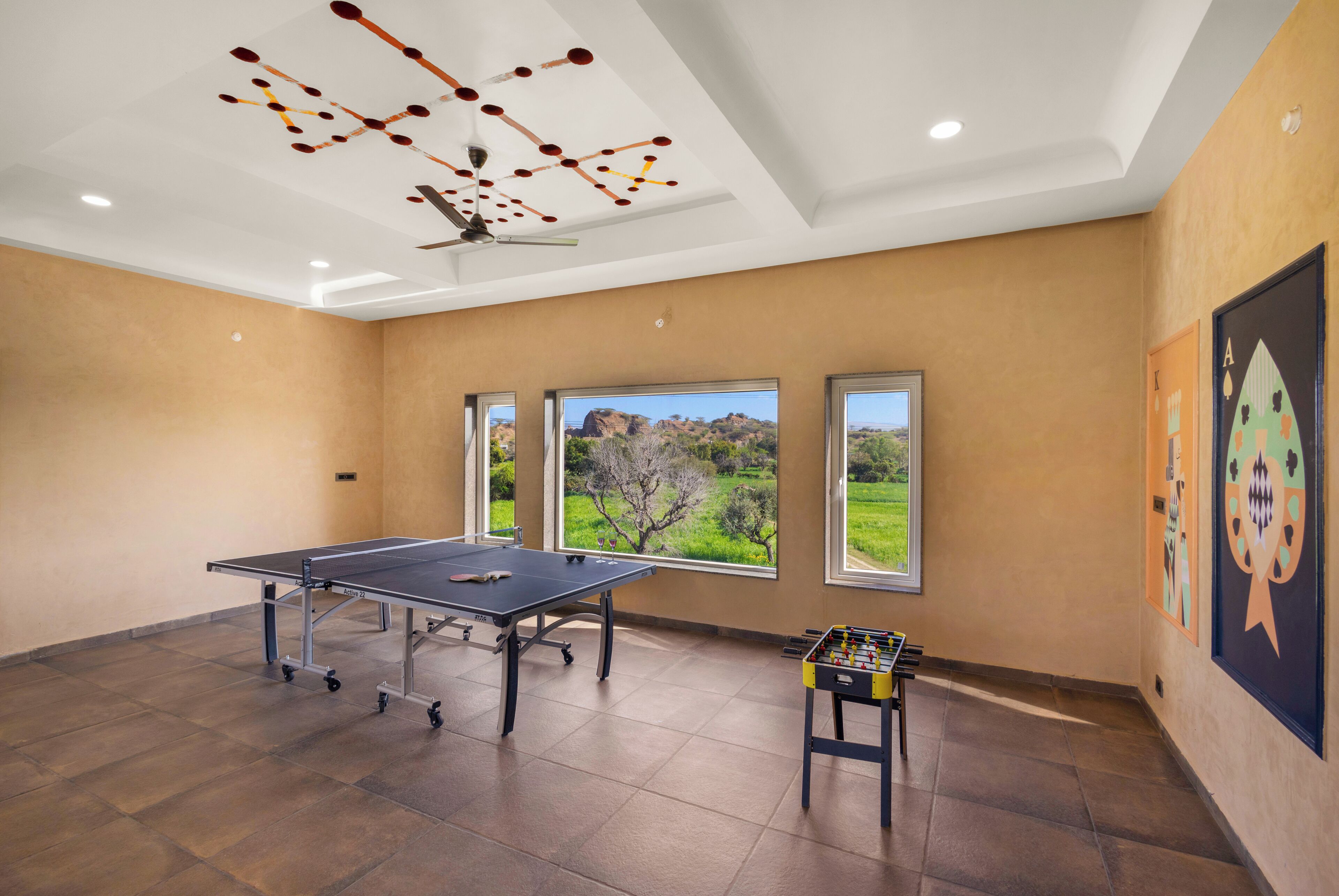 A room with a ping pong table, a foosball table, and two windows offering a view of a green landscape.