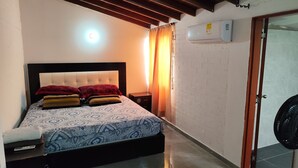A neatly made bed with patterned bedding, a bedside table with a lamp, a wall-mounted air conditioner, and a door with a fan. - Beautiful house in Cartago Valle (Cartago)