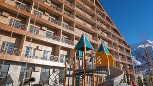 Un hotel de varios pisos con balcones, una zona de juegos infantiles y una pista de esquí al fondo.