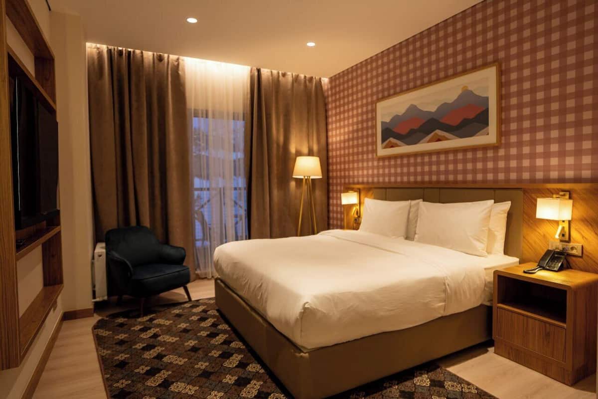 Una habitación de hotel con una cama grande, una silla, un escritorio con una lámpara, un cuadro en la pared y una ventana con cortinas.