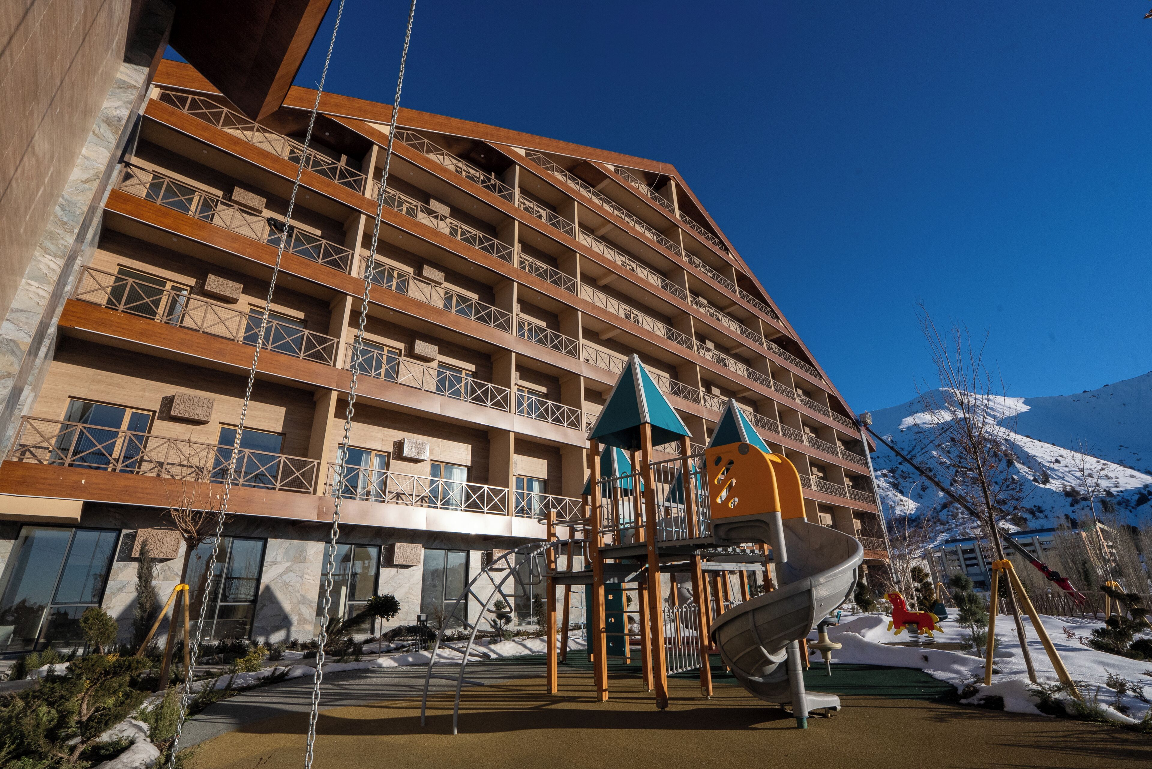 A multi-story hotel with balconies, a playground, and a snowy mountain backdrop.