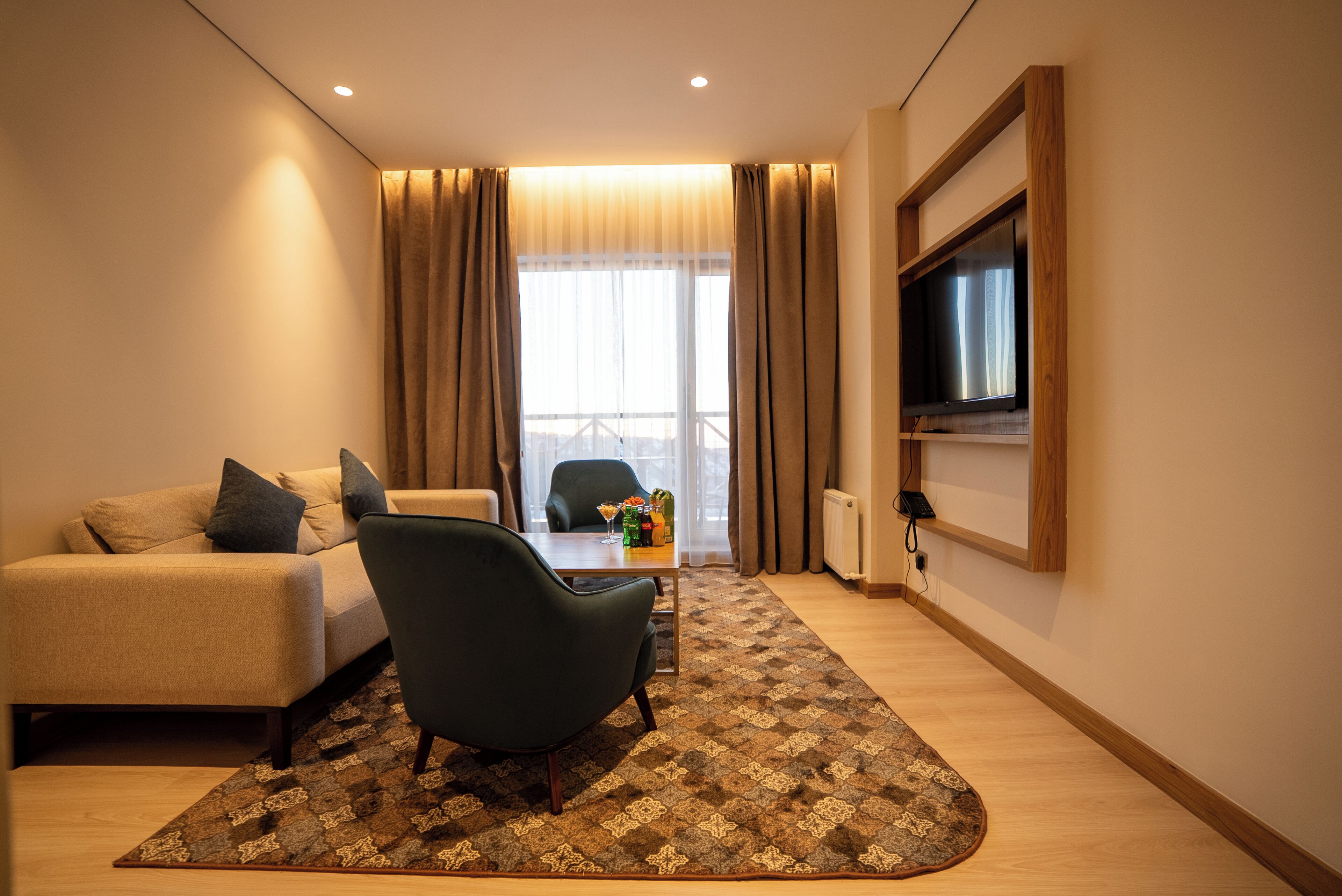 A modern hotel room with a sofa, armchair, TV, and a view of the outdoors.