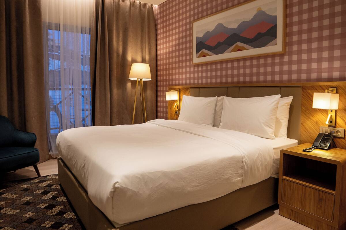 Habitación de hotel con una cama grande, un escritorio con lámpara, una silla y vistas al exterior.