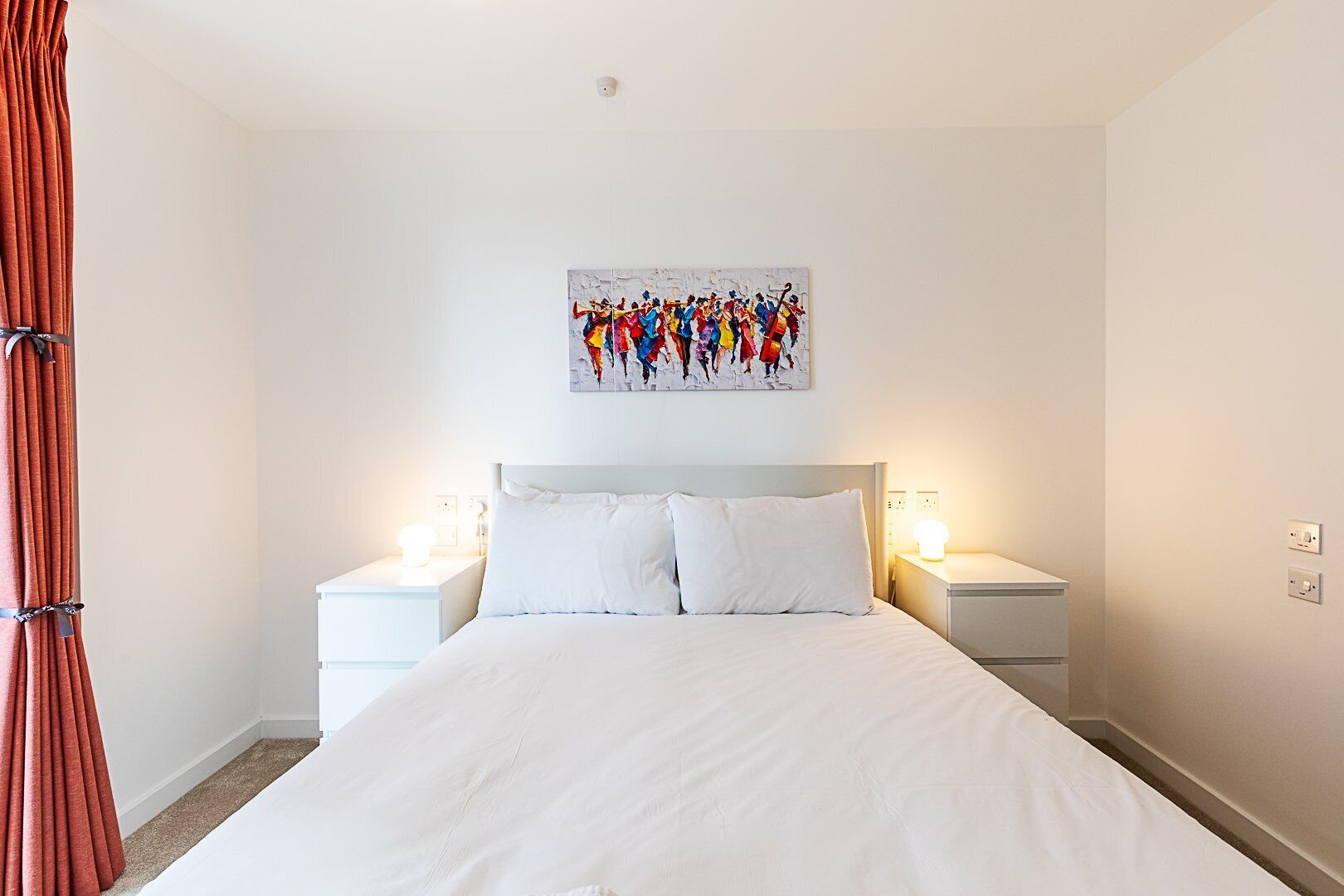 Habitación de hotel con una cama grande, dos mesitas de noche con lámparas, un cuadro en la pared y una cortina.