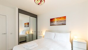 A neatly made bed with white linens and towels, a bedside table with a lamp, a large mirror, and a painting of a sunset over the ocean. - Stratford Flat near ABBA Arena (London)