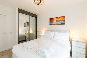 A neatly made bed with white linens and towels, a bedside table with a lamp, a large mirror, and a painting of a sunset over the ocean. - Stratford Flat near ABBA Arena (London)