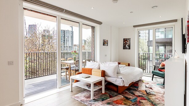 A modern bedroom with a bed, a small table, a sofa, and a balcony with a view of buildings and trees. - Stratford Flat near ABBA Arena (London)