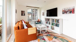 A modern living room with a sofa, a coffee table, a bookshelf, and a TV mounted on the wall. - Stratford Flat near ABBA Arena (London)