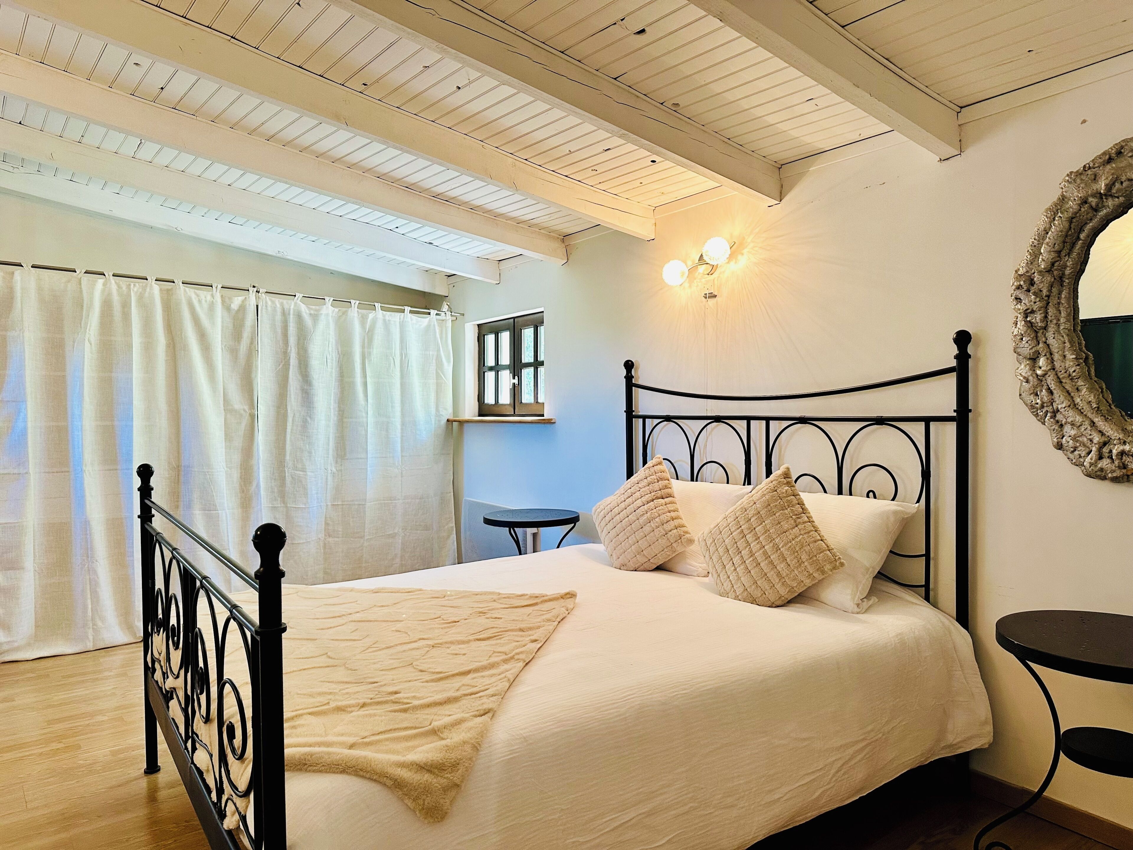 A bedroom with a bed, a window with curtains, a wall-mounted light fixture, and a round mirror.