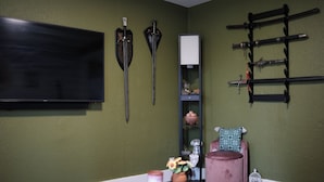 Uma sala de estar com televisão de tela plana, espadas montadas, suporte para espadas, uma cadeira rosa, uma mesinha com uma vela e um vaso com flores.