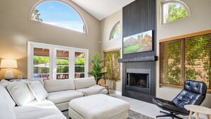 A modern living room with a sectional sofa, a fireplace, and large windows with views of greenery. - Bright Phoenix Oasis w/Private Pool & Mtn Views (Phoenix)
