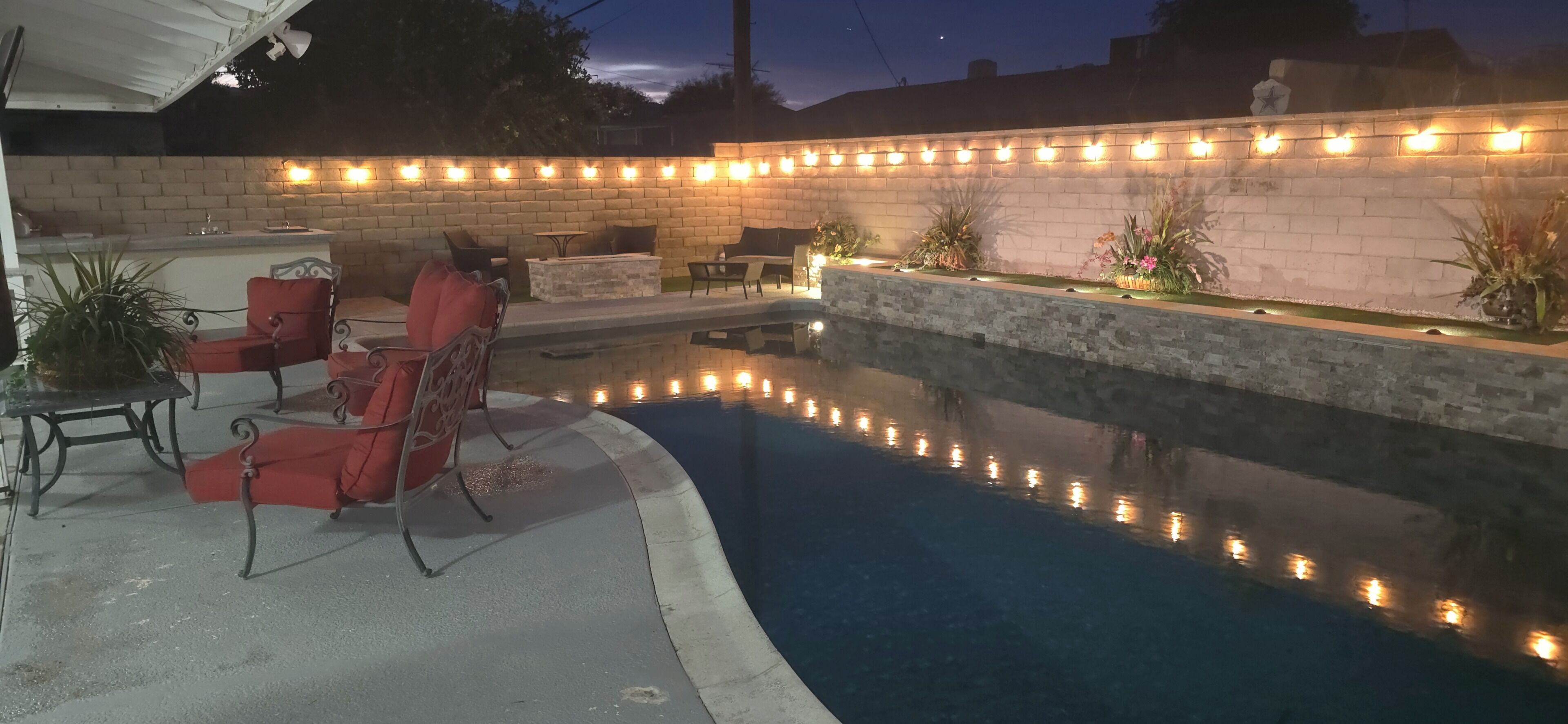 A swimming pool with a brick wall and string lights, surrounded by outdoor furniture and potted plants.