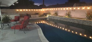 A swimming pool with a brick wall and string lights, surrounded by outdoor furniture and potted plants. - Beautiful home close to Coachella  Stagecoach, PGA west, and The Thermal club. (Thermal)