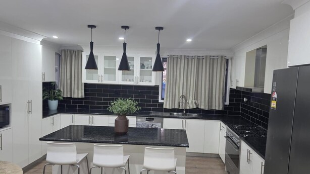 A modern kitchen with white cabinets, black countertops, a central island with chairs, pendant lights, and a refrigerator.
