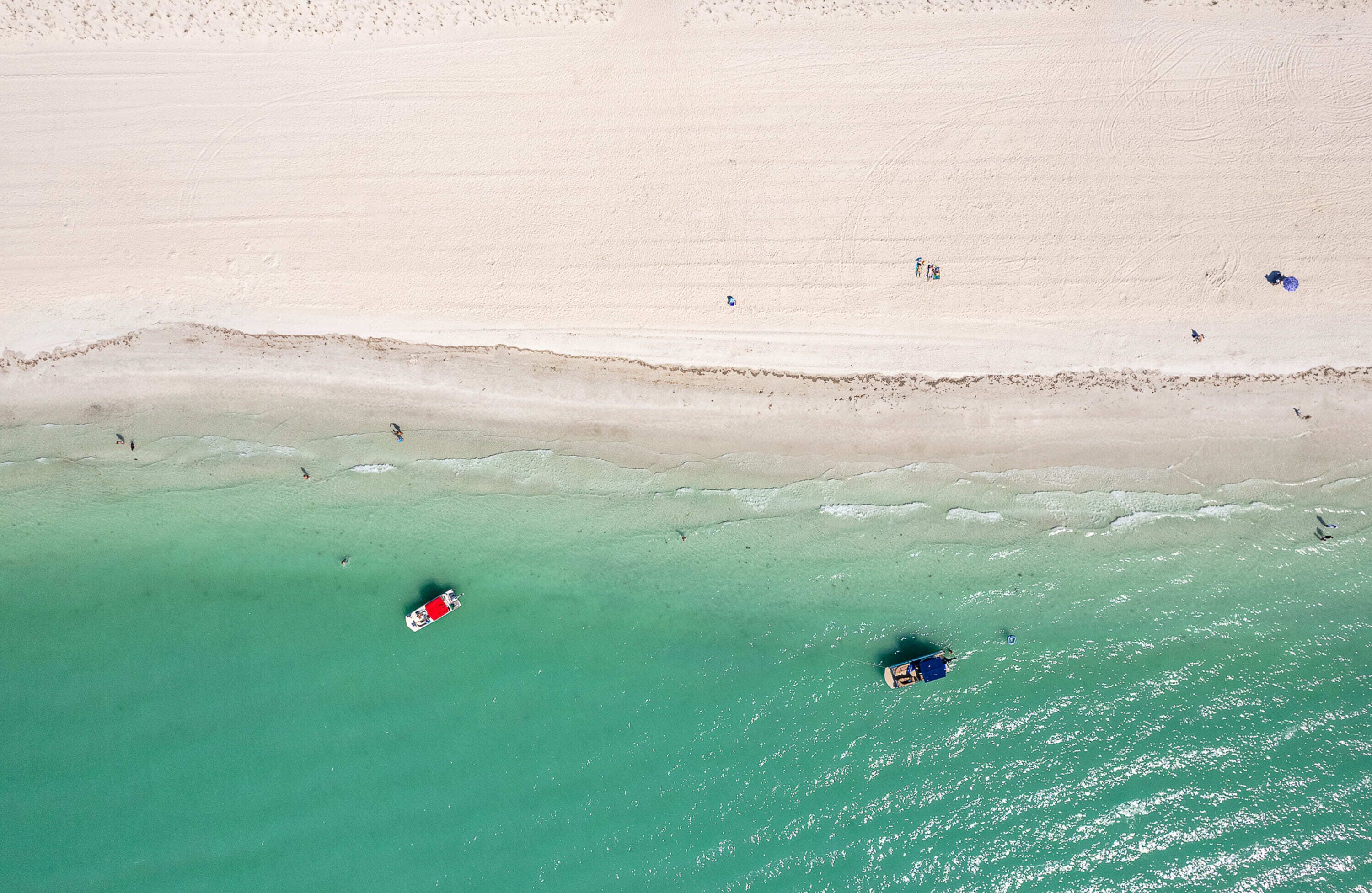 Oversigtsbillede af en strand med klart turkis vand, en sandet kystlinje og et par personer og både.