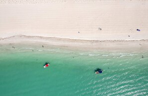 Ilmakuva rannasta, jossa on kirkas turkoosinvärinen vesi, hiekkainen ranta sekä muutama ihminen ja vene.