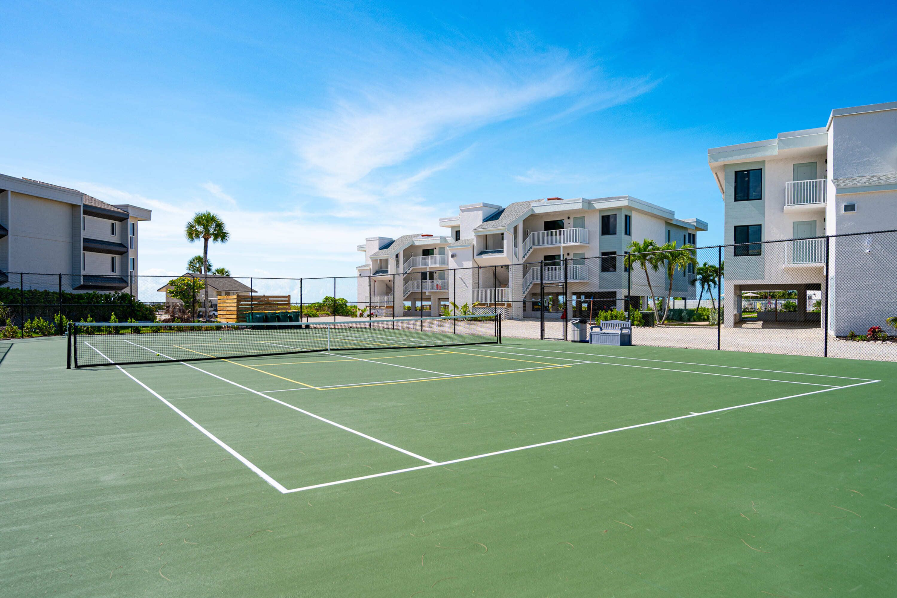 A tennis court with a net, surrounded by a fence, and adjacent to multi-story buildings.