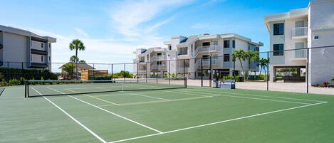 A tennis court with a net, surrounded by a fence, and adjacent to multi-story buildings.