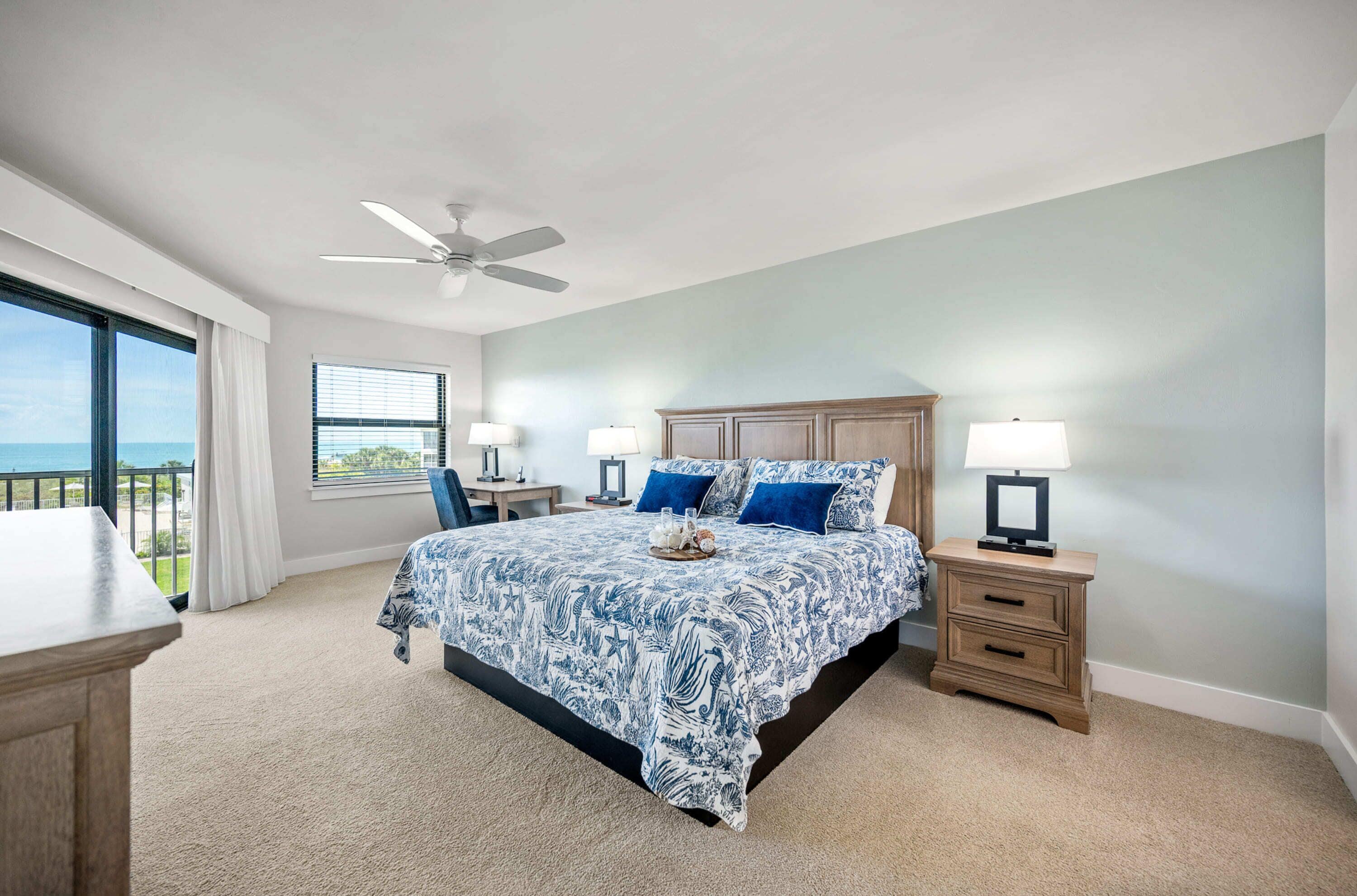 A bedroom with a bed, two bedside tables with lamps, a ceiling fan, a window with curtains, and a view of the ocean.