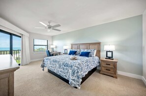 A bedroom with a bed, two bedside tables with lamps, a ceiling fan, a window with curtains, and a view of the ocean.