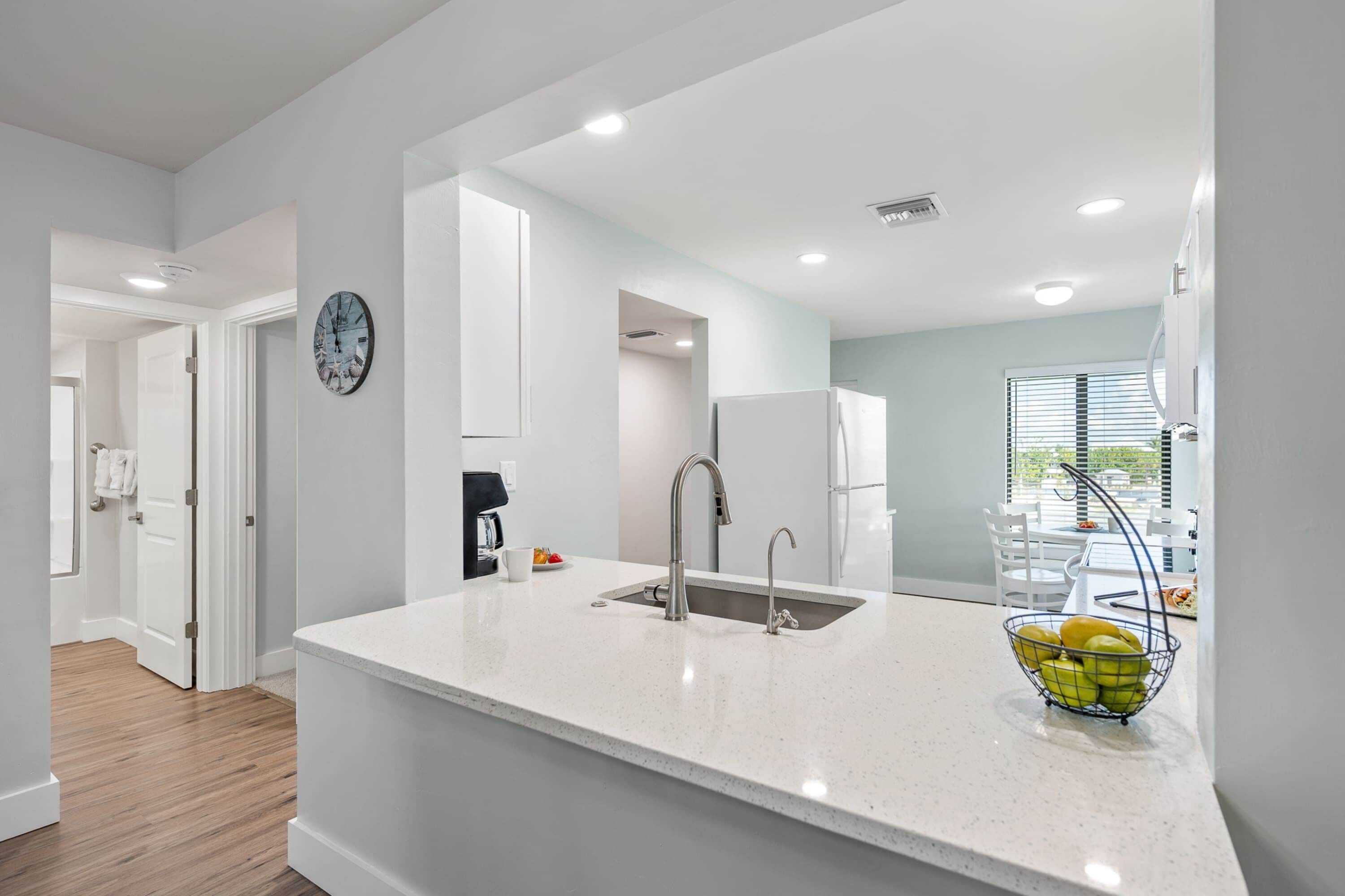 A modern kitchen with a white countertop, stainless steel sink, and a fruit bowl.
