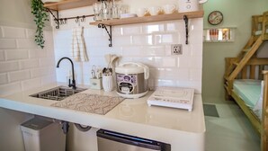Una cocina con un lavamanos, una cocedora de arroz, una balanza y un estante de madera con vajilla.