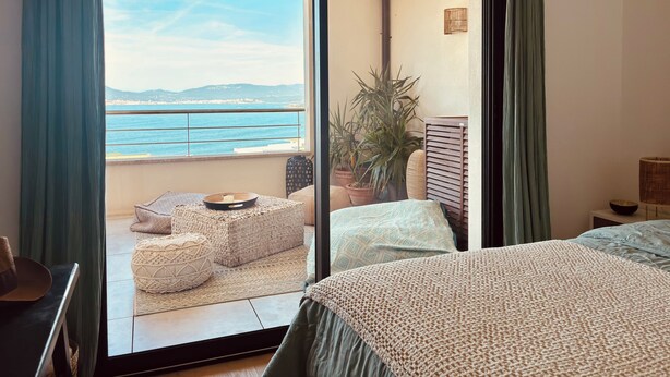 Una habitación con cama, vista al mar, balcón con asientos y una planta en maceta.