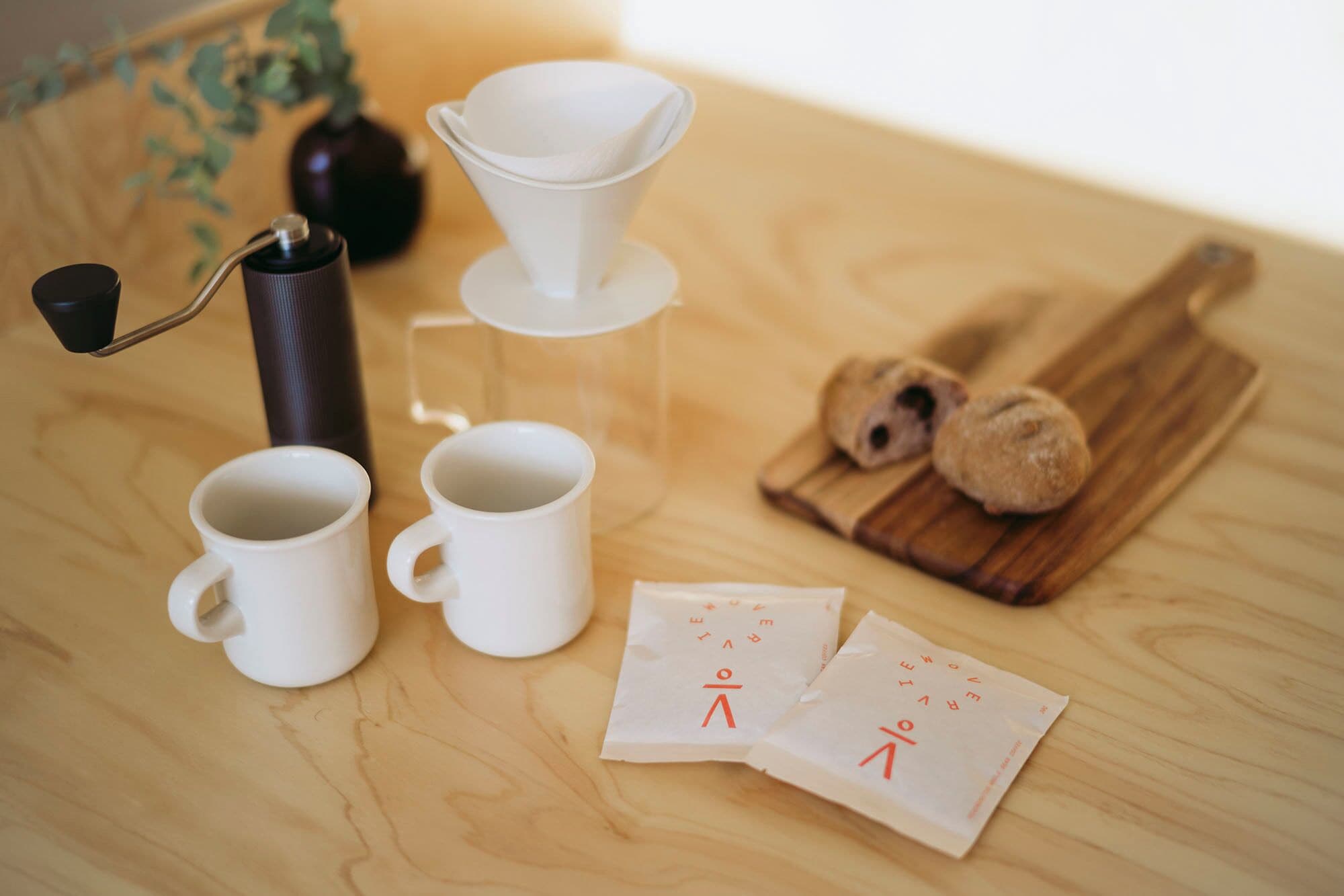 A wooden table with white cups, a coffee maker, a wooden board with cookies, and two napkins with a logo.