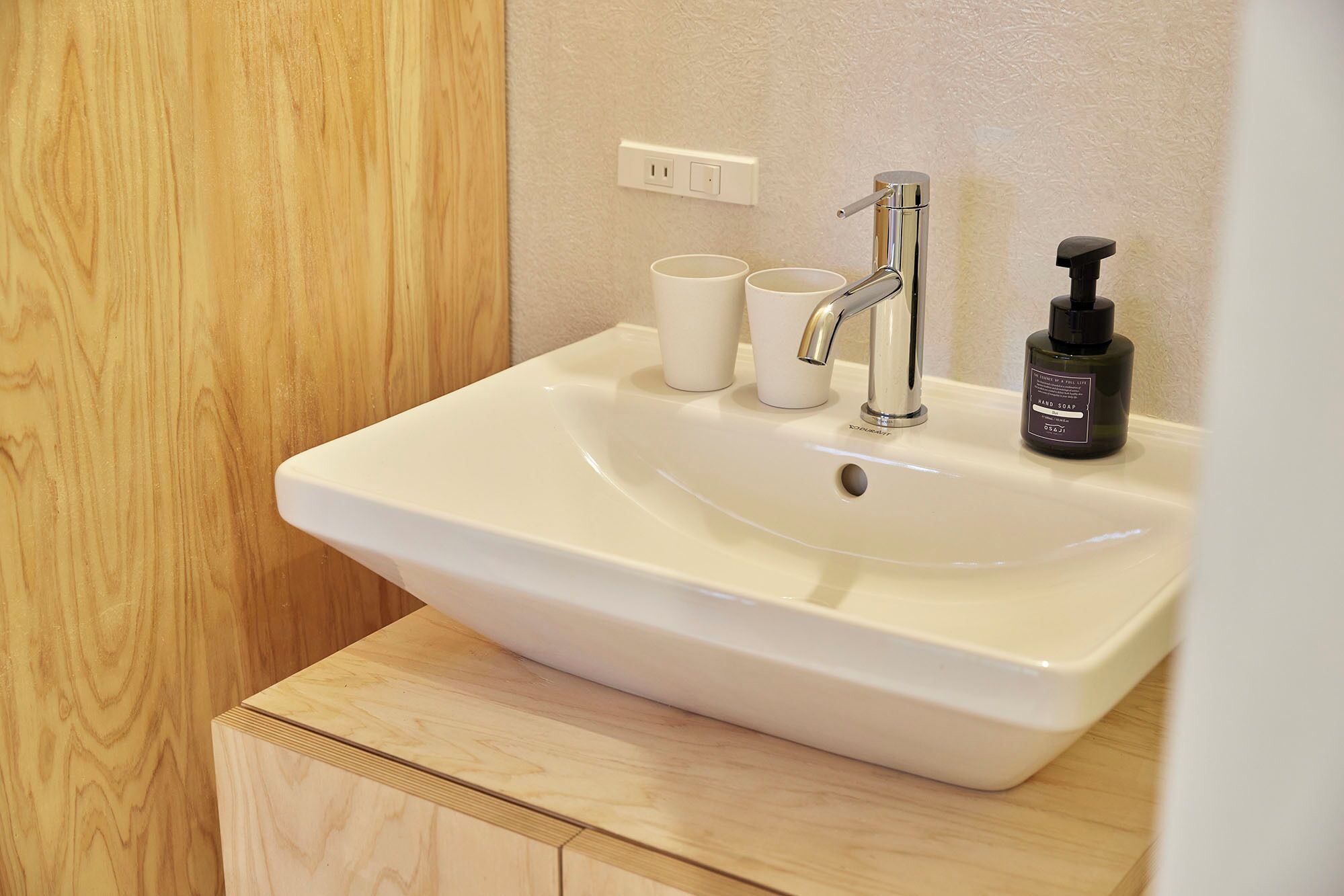 A bathroom with a white sink, a faucet, two white cups, and a black soap dispenser.