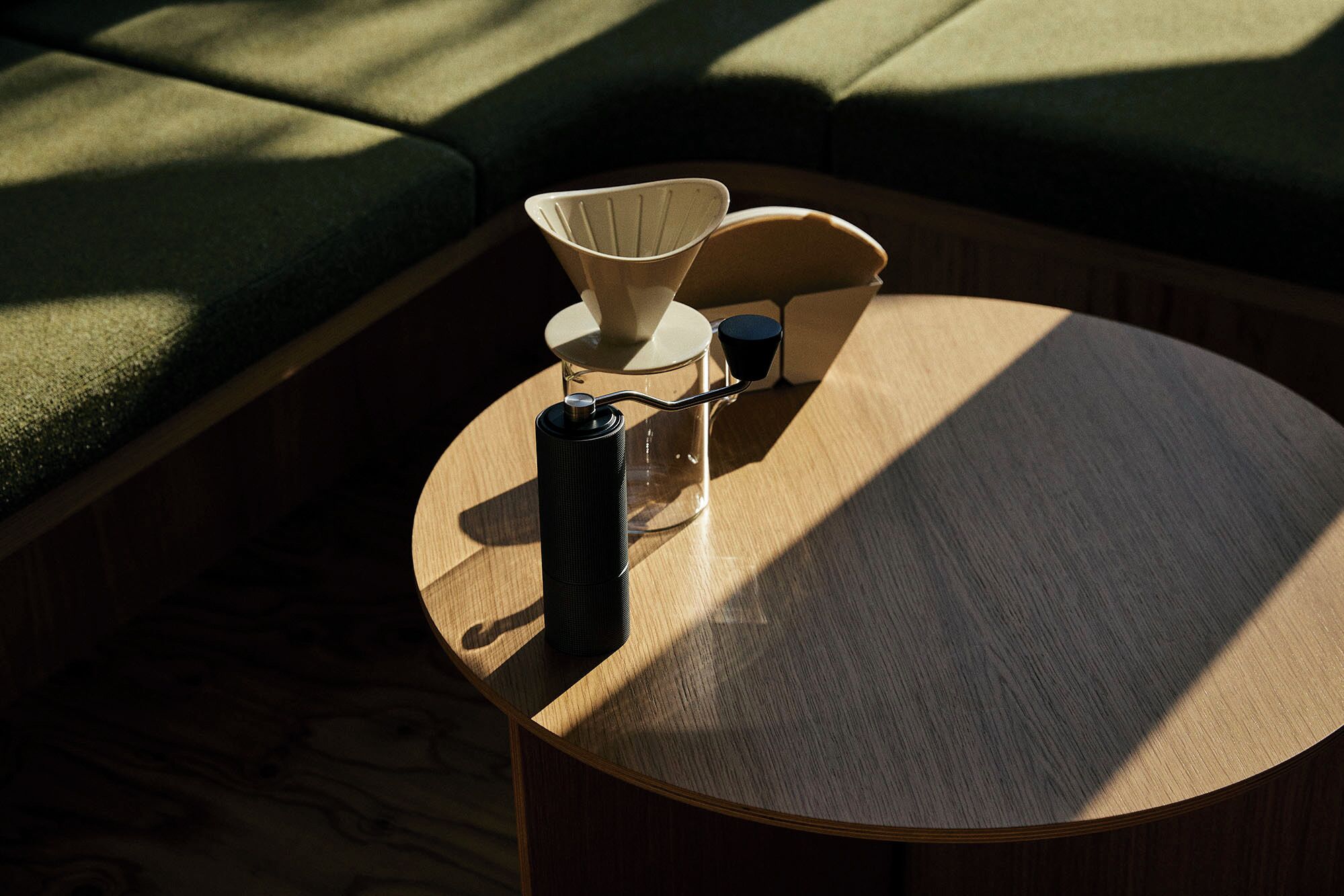 A round wooden table with a kettle and a cup on it, placed on a wooden floor with a green sofa in the background.